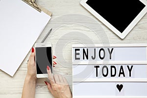 Work space with tablet, smartphone, notepad and `Enjoy today` word on lightbox over white wooden background, top view. Female ha