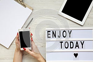 Work space with tablet, smartphone, notepad and `Enjoy today` word on lightbox over white wooden background, top view. Female ha