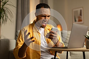 Work stress concept. Angry young black man shouting into smartphone, working online on laptop pc at home office