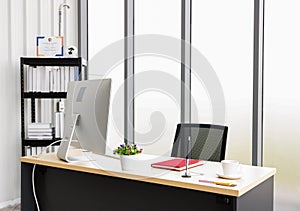 The work table is located in the office room. There were computers and red notebooks on the table. There is a bookshelf in the
