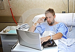 Workaholic business man in hospital room lying in bed sick and injured working with mobile phone computer laptop photo