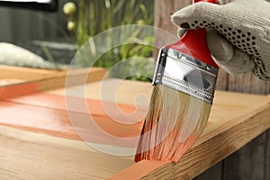 Worker applying coral paint onto wooden surface against blurred background, closeup. Space for text