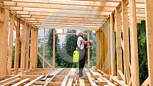 Worker applying fire retardant using sprayer, while constructing wooden frame house near forest