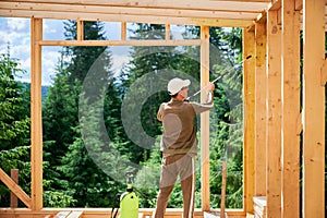 Worker applying fire retardant using sprayer, while constructing wooden frame house near forest