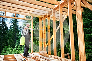 Worker applying fire retardant using sprayer, while constructing wooden frame house near forest photo