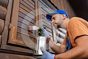 Worker applying protective varnish or wood oil on wooden house cottage exterior walls and window shutters
