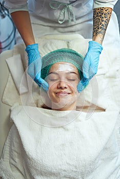 Worker of beauty salon pleasantly massages with rub cream face of woman on couch. Cosmetological procedure. photo