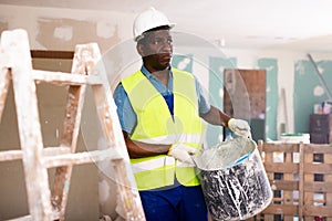 Worker in blue overalls and hardhat dragging various painting tools in room being renovated