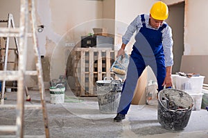 Worker in blue overalls and hardhat dragging various painting tools in room being renovated