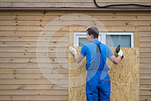 A worker in a blue uniform, blocks the window of the house with a protective shield made of wood, from thieves, when moving to