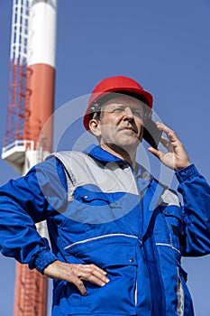 Worker In Blue Uniform And Red Hardhat Talking On Smartphone While Standing In Front Of Power Plant Chimneys