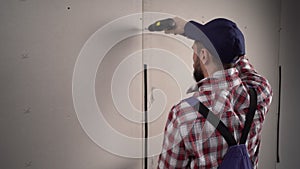 Worker builder in blue overalls installs plasterboard drywall at a construction uses electric screwdriver.