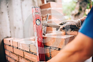 Worker building exterior walls, using hammer and level for laying bricks in cement. Detail of worker with tools