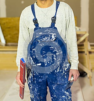 Worker in dirty uniform or boilersuit with water level and palette-knife in his hands in the room of apartment that is under