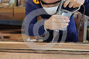 Worker drills a hole using electric drill machine in carpentry workshop. He is wearing safety equipment .