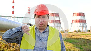 Worker or engineer looking directly at the camera and showing thumb down standing in front of a power station