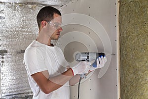 Worker in goggles with screwdriver working on insulation. Drywall on wall beams, insulating rock wool staff in wooden frame.