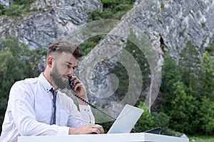 worker guy at desk using computer laptop, phone, looking at screen. Outdoor young hipster having conversation on phone