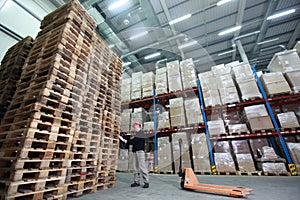 Worker with hand pallet truck at large stack of wooden pallets in storehouse