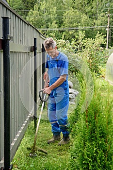 Worker with a hand trimmer in his hands mows the grass in front of the house. The trimmer is in the hands of a man. The