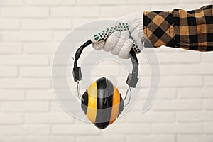 Worker holding safety headphones against white brick wall, closeup. Hearing protection device