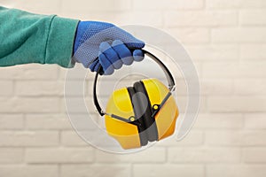 Worker holding safety headphones against white brick wall, closeup. Hearing protection device