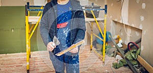 Worker holds a palette-knife for flattening the ceiling from wooden platform in room of apartment is inder construction