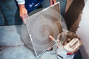 Worker installing bathroom ceramic floor tiles, adding flexible cement adhesive with comb trowel