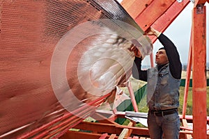 The worker insulates the attic of the house with a hydrofilm, secures the insulation with the help of a stapler