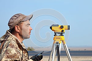 Worker with level, surveyor builder with geodesy equipment close to highway, with mobile phone, smiling.