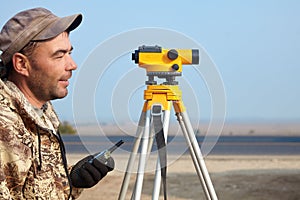 Worker with level, surveyor builder with geodesy equipment close to highway, with mobile phone, smiling.