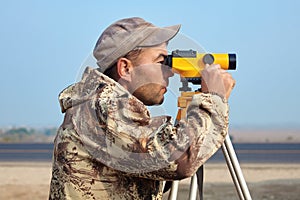 Worker with level, surveyor builder with geodesy equipment close to highway, with mobile phone, smiling.