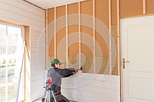 The worker makes finishing works of walls with a white wooden board, using laser line level. Building heat-insulating