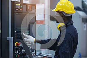 Worker man with yellow helmet and ear protection typing keyboard of laptop computer at factory