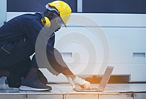 Worker man with yellow helmet and ear protection typing keyboard of laptop computer at factory