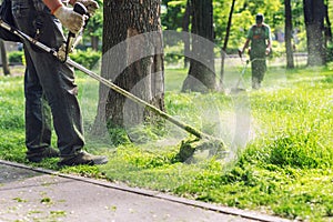 Worker mowing tall grass with electric or petrol lawn trimmer in city park or backyard. Gardening care tools and equipment.