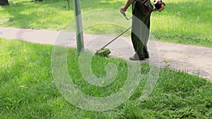 Worker mowing tall weed grass with electric or petrol lawn trimmer in city park or backyard. Gardening care tools and