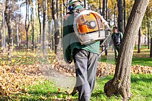 Worker operating heavy duty leaf blower in city park. Removing fallen leaves in autumn. Leaves swirling up. Foliage