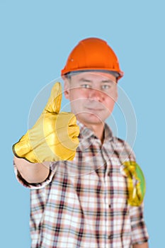 A worker in an orange helmet and shirt shows a thumbs up in yellow gloves. Selective focus