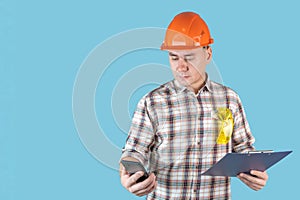 A worker in an orange helmet writes text messages holding a phone and clipboard with papers on a blue background