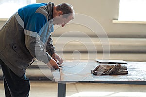 Worker in overalls works in workshop at workbench. Man marks metal sheet. Authentic portrait of worker at work