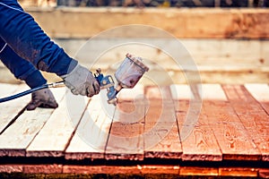 Worker painting brown timber, renovating exterior wooden fence. Worker using spray gun