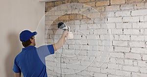 Worker painting white brick wall with electric paint sprayer