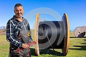 A worker with pliers stands in front of a meadow with a large cable reel.