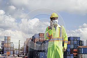 Worker with protection face mask wearing safety clothing and helmet standing and talking on mobile phone at logistic cargo