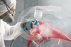 Worker in protective mask and jumpsuit overall spraying base coat of paint on car body elements in painted chamber using