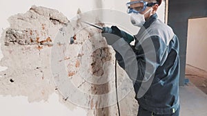 Worker in protective suit demolishes plaster wall. Dirty, hard work. Personal protective equipment. Helmet, respirator