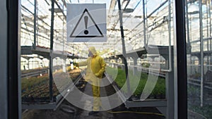 Worker in protective uniform hosing plants in greenhouse, warning sign on door