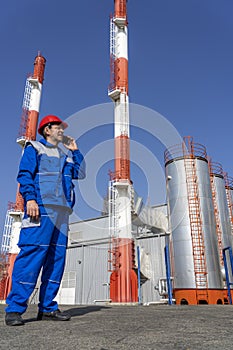 Worker in Red Hardhat and Blue Uniform Talking on Mobile Phone and Standing Against Power Plant Chimneys and Blue Sky Background