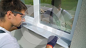 Worker in safety glasses and protective gloves installing metal sill on external PVC window frame